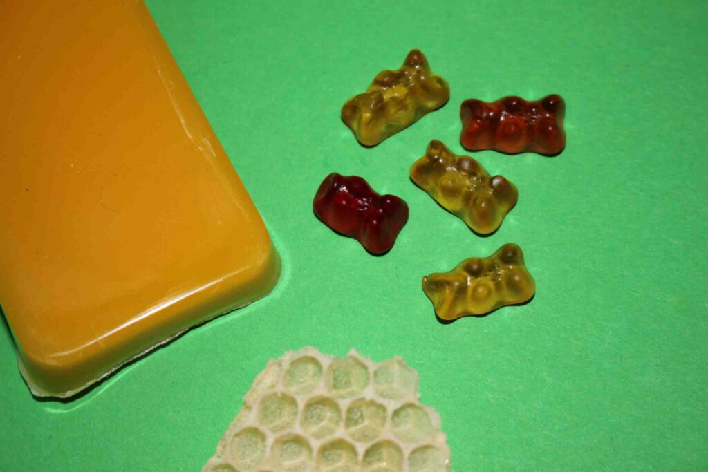 Block aus Bienenwachs, ein Stück Bienenwabe und Gummibärchen aus Bienenwachs auf grünem Hintergrund.